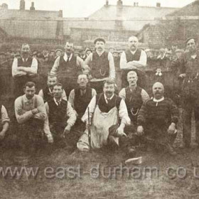 A group of middle-aged men in front of goal posts and a large crowd. The man at centre is holding a small rifle.
Do you have any idea what is going on, where or when the photograph was taken??