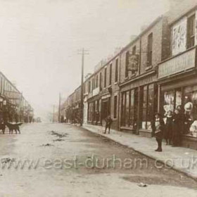 Market Street, Hetton Downs, c 1910. The young lad outside the sewing machine shop appears to be holding what my father always called an "ollygig" (presumably a corruption of "whirlygig").  (We called them "boolers" in Seaham, DA) This simple toy consisted of a steel ring or hoop (often from a wooden barrel) with a hook by means of which the boy would keep it in motion and guide it whilst running alongside it; part of the attraction seems to have been the noise it made!   Caption by Sheldon Clark