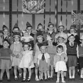 Coronation Day Party at the Miners' Hall in South Hetton in June 1953.  My sister, Lesley Clark, is standing on her own, right at the front, whereas I believe it is my face that can just be made out at the right hand end of the back row, partially obscured by the paper hat worn by Kenneth Penfold.  Standing two places to Kenneth's left (at the right hand end of the middle row) is, I believe, Robert Nesbitt.  Two places to my right (as seen from my place on the stage) is, I think, Eric Robinson.  Eric was the undisputed, if self-appointed, leader of our "gang", which was not as sinister as it might at first appear; in our games in the farmer's field, Eric took on the roles of every villain or monster whilst simultaneously managing to lead our band of explorers or spacemen or cowboys through every hazard - just like John Wayne.  The boy at the far left of the back row could be Dennis Smithson, but I'm not at all sure.  The name of the boy at the far left of the middle row could be Alan Bainbridge.  Standing between Eric Robinson and me is, I think, Raymond Jones.           .Photograph and caption from Sheldon Clark.