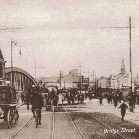 A view over the old Stephenson Bridge towards North Bridge Street. The building on the right beneath the church steeple is the Monkwearmouth Picture Hall opened in 1906 by George Black and was established in an old chapel. It later became the Bridge Cinema in 1916 and then the Bromarsh in 1919. It was destroyed by enemy bombs in 1943. The drays, left and centre were hauled by Percheron horses belonging to Vaux Breweries. This also formed part of the route of the first horsedrawn trams from Roker to Fawcett Street, commencing in 1879. One of the new electric trams can be seen crossing the bridge, middle right.