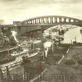 Bridges and  coal drops on the Wear.  c1920 ?
In the 1700's coal was being taken to river staiths on horse drawn wagonways to be shipped to London. At the staiths the coal was manhanded  on to small keels from which it was loaded to the ship. By the 1800's with the advent of the first steam locomotives the steel rails were extended over the river so that the coal could either be sent down a shoot into the ship or the waggon could be lifted over and the coal dropped in. This photo shows the drop system at the Wear with the lifts clearly visible but  some doubt must be expressed about the passing ship which appears to be going downriver at high speed and without the aid of tugs. 
Information from Len Charlton.